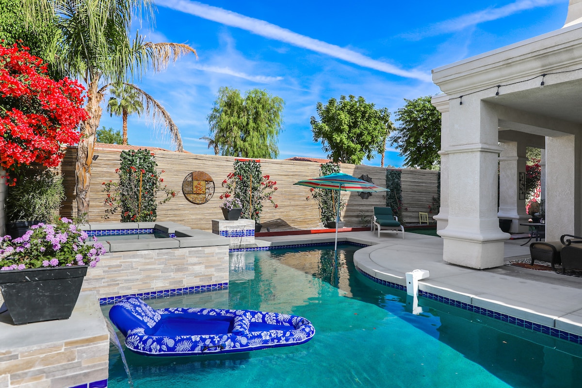 The image features a well-maintained swimming pool surrounded by vibrant greenery and floral arrangements. A spacious deck is equipped with lounge chairs, an umbrella, and a decorative floating device. The clear water reflects the blue sky, creating a serene outdoor atmosphere.