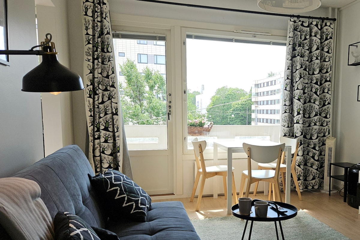 A bright living area features a comfortable gray sofa adorned with cushions. A small dining table with light chairs is positioned near large windows that offer views of the surrounding greenery. Elegant curtains frame the windows, allowing natural light to fill the space.