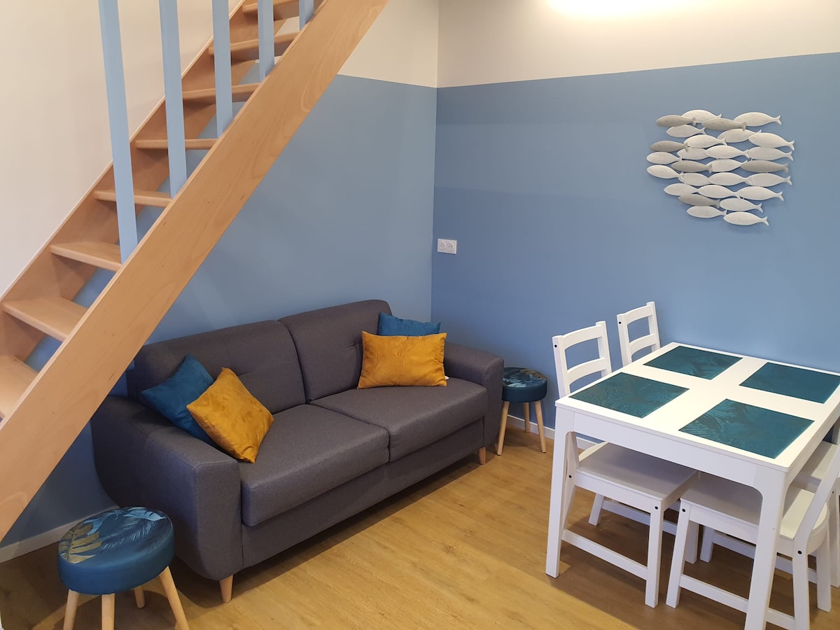 A cozy living area features a gray sofa adorned with bright cushions and a white dining table with teal accents. Light-colored wooden floors contribute to a welcoming atmosphere, while a wooden staircase leads to the upper level. Decorative fish art is mounted on the wall.