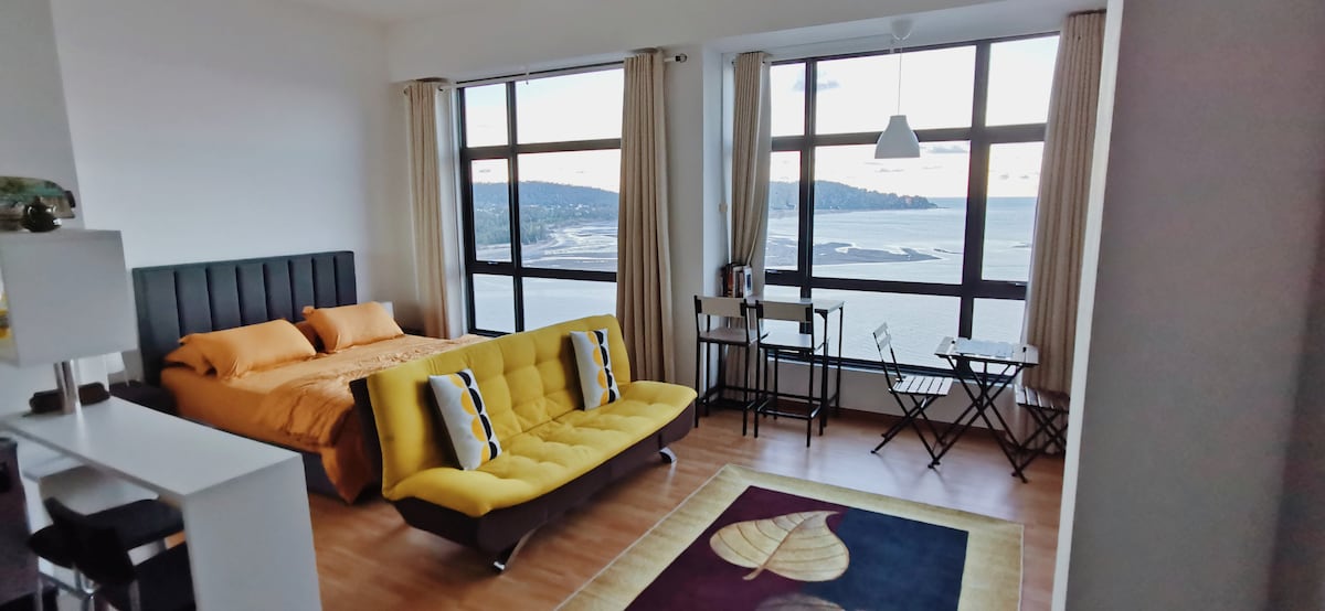 A bright and open studio space is featured with large windows overlooking the sea. A spacious orange bed is complemented by a yellow sofa bed. A small dining area with two chairs is positioned near the windows, while decorative elements include a patterned rug on the wooden floor.