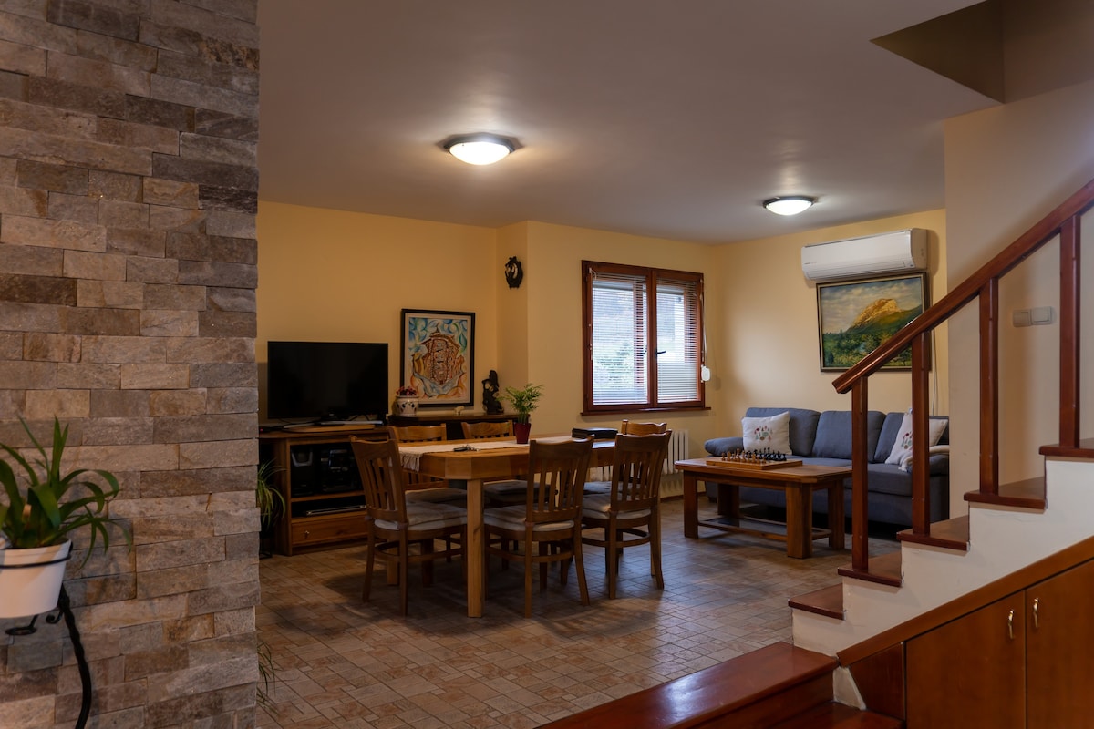 A spacious living area features a wooden dining table surrounded by chairs, with a comfortable sofa positioned nearby. A flat-screen television is mounted on the wall, and large windows allow natural light to fill the room. Decorative artwork is displayed, adding character to the space.