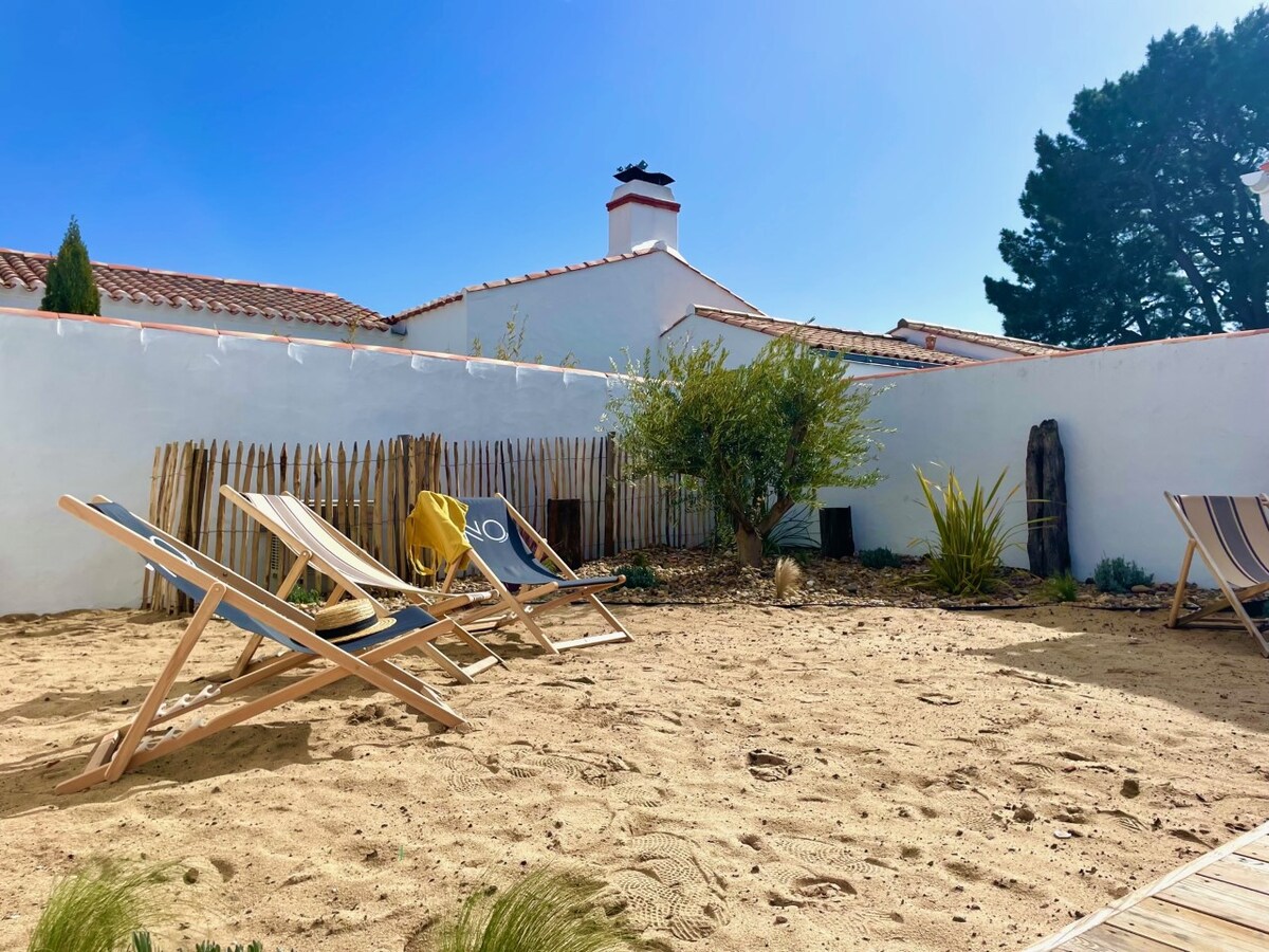 A sandy garden area is presented, featuring several lounge chairs positioned for relaxation. Lush greenery, including a small tree and shrubs, adds a touch of nature. A white wall surrounds the yard, enhancing privacy, while the sun illuminates the space, creating a welcoming outdoor atmosphere.