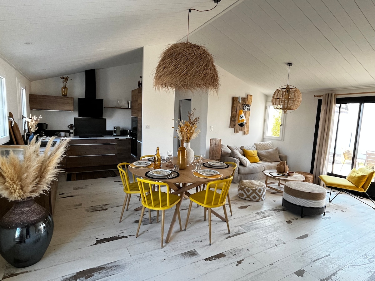 A bright living area features a stylish dining table with yellow chairs, set for six. A comfortable sofa and decorative poufs are arranged near large glass doors that open to the terrace. Natural elements, including dried plants and light fixtures, add warmth to the space.