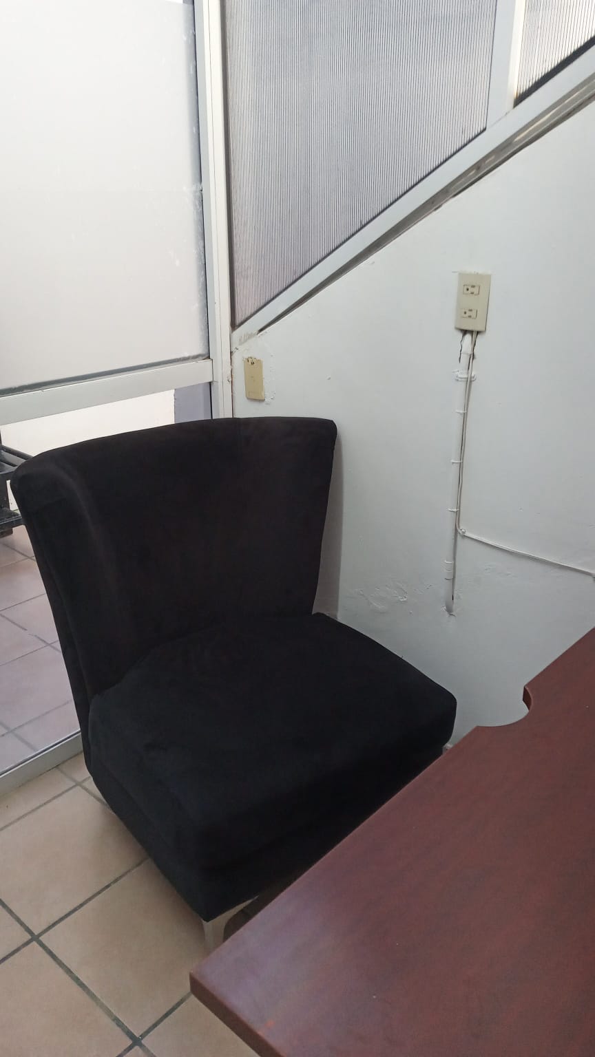 A black upholstered chair is positioned near a wall, next to a wooden desk. The chair's curved back and cushioned seat provide comfort. Natural light filters through a nearby glass door, illuminating the simple yet functional workspace.