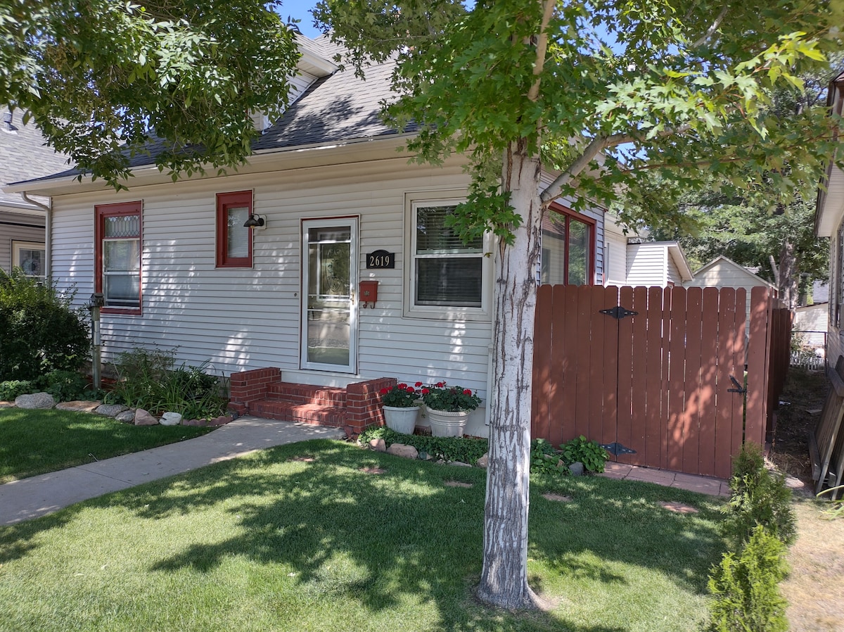 The charming exterior of the Rancher's Lodge features a welcoming entry with a red door framed by windows. Flower beds with colorful blooms add a touch of nature, while a well-maintained lawn and privacy fence enhance the inviting atmosphere.