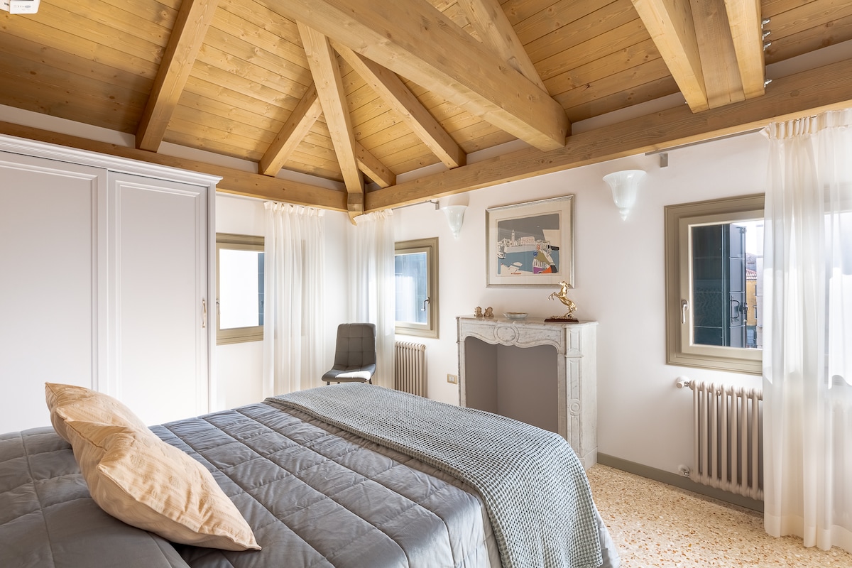 A spacious bedroom features a high wooden ceiling and warm light streaming through large windows. A comfortable bed is adorned with soft blankets, while an elegant fireplace adds a touch of charm to the space. A minimalist chair is placed near the window.