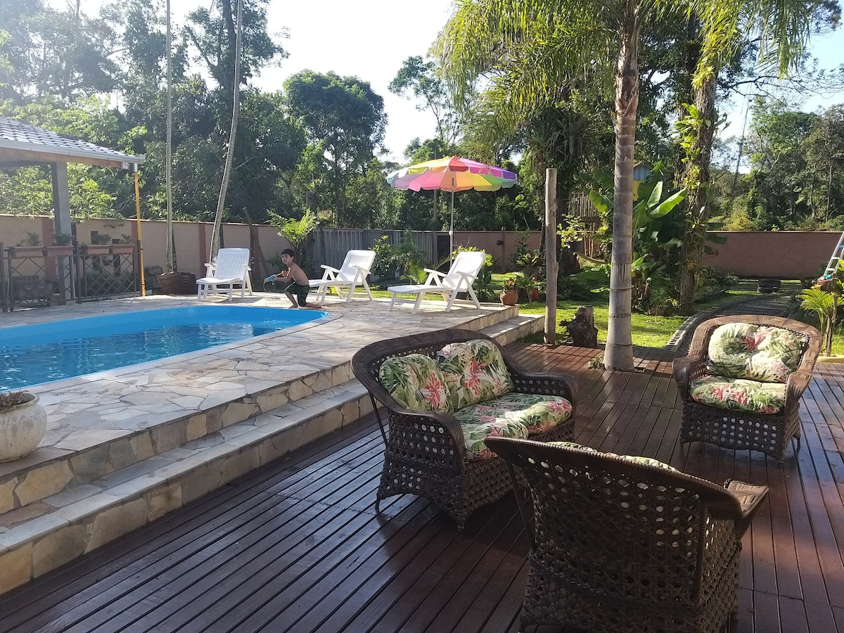 A well-maintained outdoor area features a stone-lined pool surrounded by lounge chairs. A vibrant umbrella offers shade, while potted plants and palm trees enhance the greenery. Comfortable seating is provided with wicker chairs adorned with floral cushions on a wooden deck.