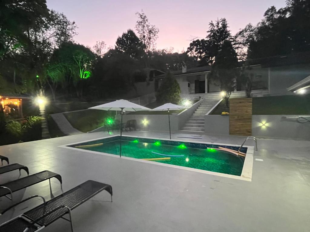 An outdoor area features a well-lit swimming pool with green lighting, surrounded by lounge chairs. Two umbrellas provide shade, while steps lead up to a landscaped area and the property. The serene environment is enhanced by the evening glow of the surrounding trees.