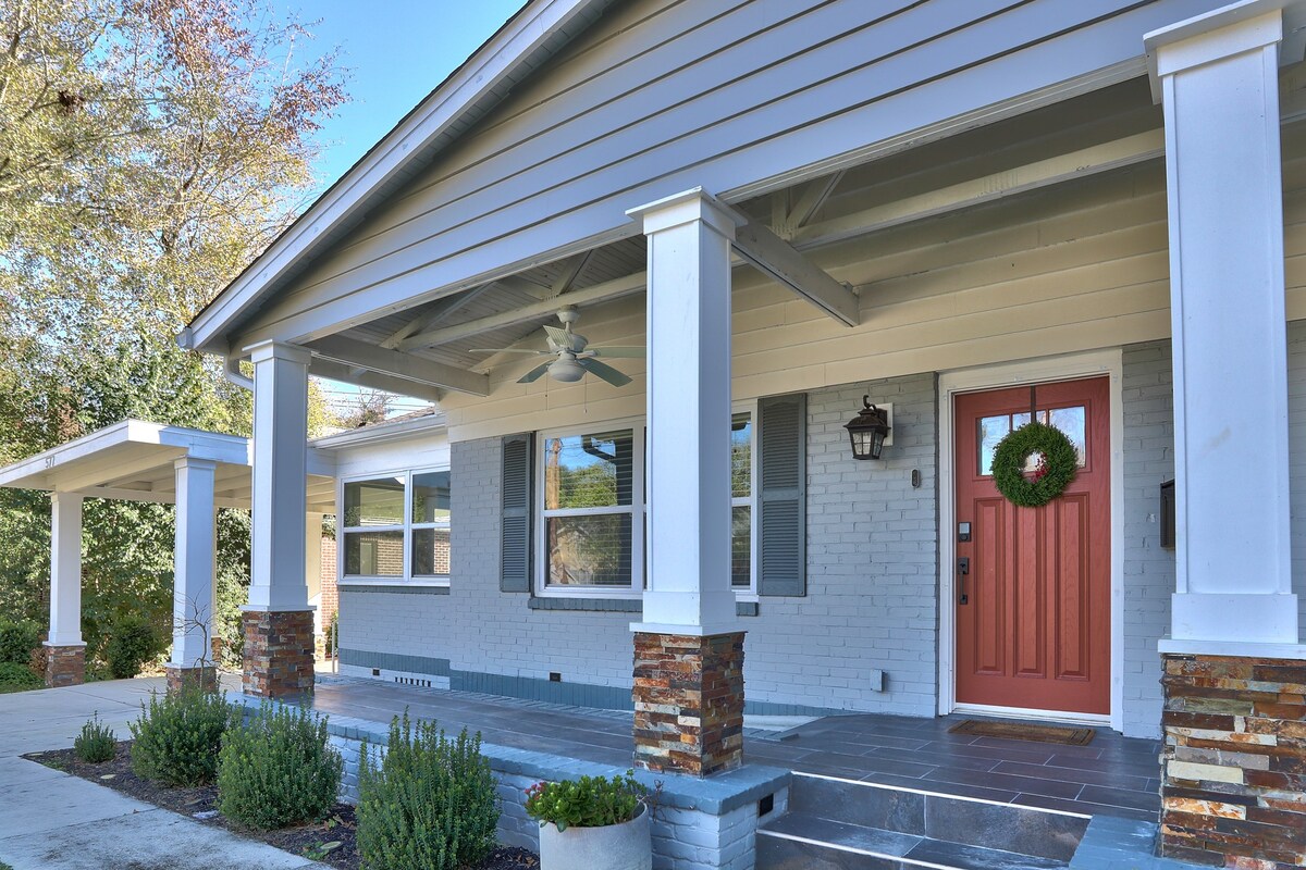 The front porch features a welcoming entrance with a stylish red door adorned with a wreath. Supportive columns and a fan enhance the shaded area. Stone accents and well-maintained greenery line the porch, contributing to an inviting outdoor space.