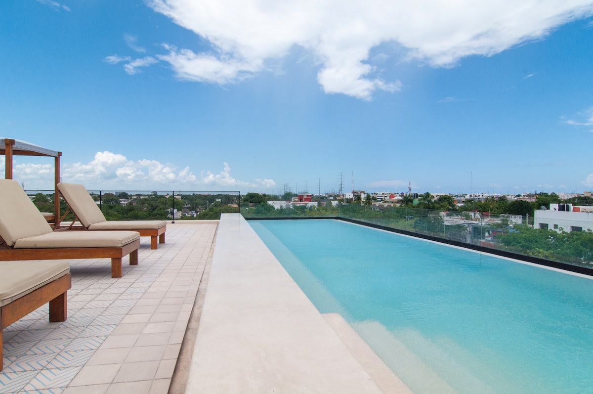 A modern rooftop infinity pool stretches across the image, offering a refreshing view over the cityscape. Two lounge chairs are positioned on the nearby deck, inviting relaxation under the clear blue sky.