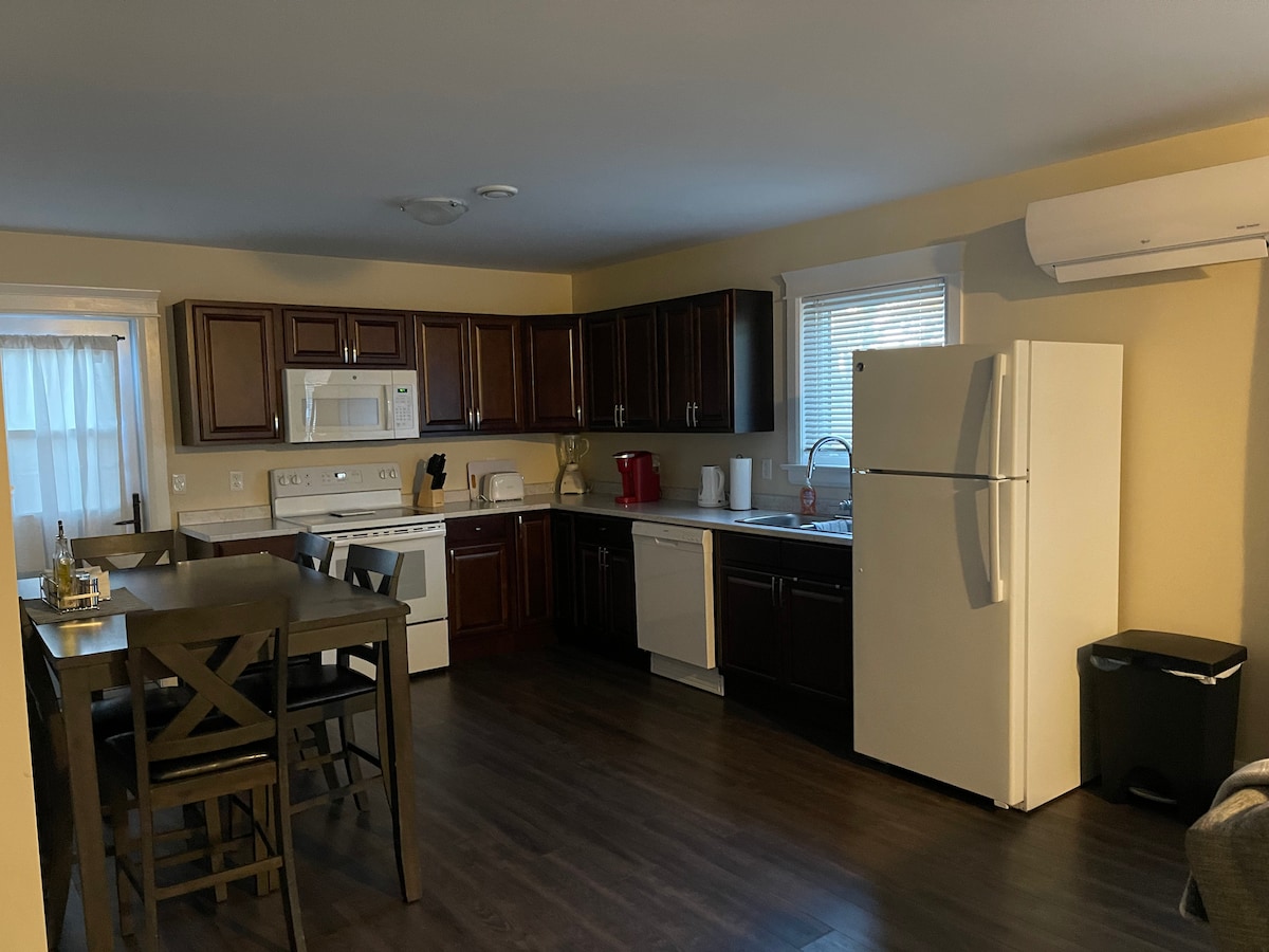 The kitchen features dark wood cabinets and modern appliances, including a refrigerator, oven, and microwave. A small dining table with four chairs is positioned near the window, providing natural light. The space has wood-like flooring and an air conditioning unit mounted on the wall.