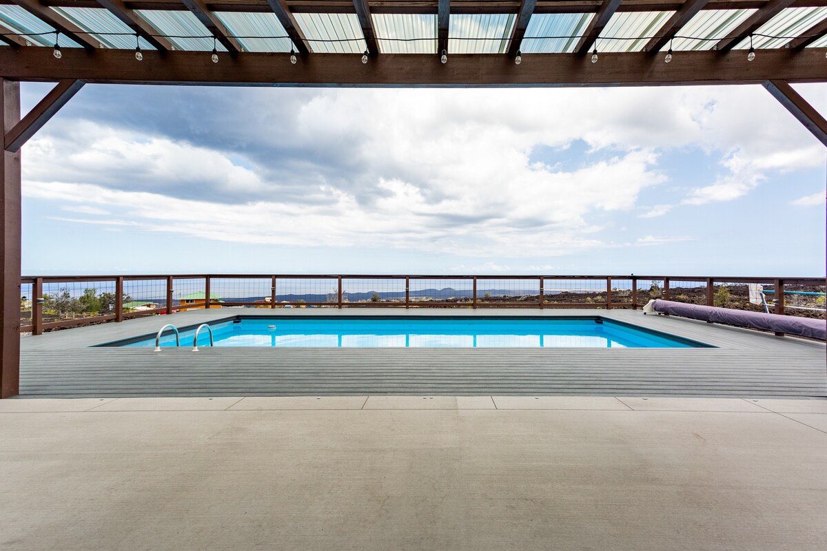 An outdoor pool area features a turquoise pool set against a backdrop of ocean views and cloudy skies. A spacious deck surrounds the pool, with wooden railings providing an unobstructed view. The pergola overhead adds shade, enhancing the inviting atmosphere of this outdoor retreat.