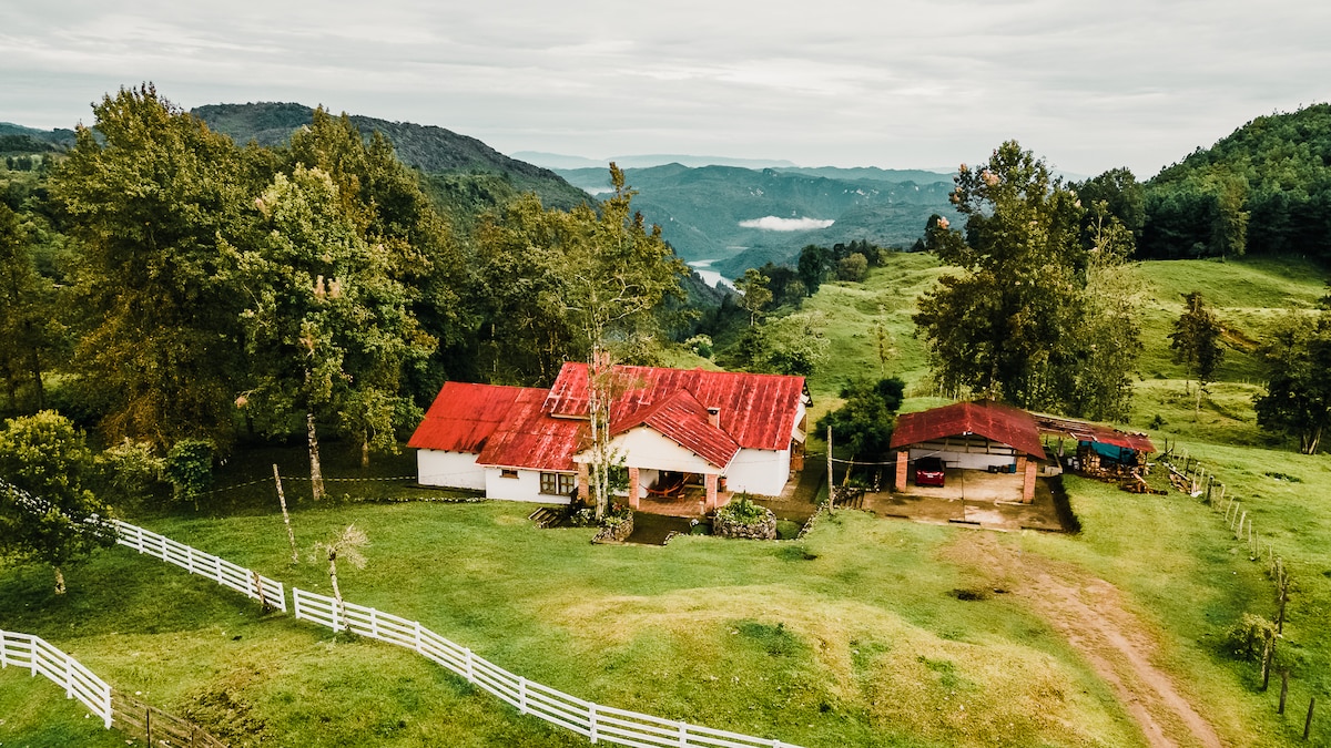 A scenic view showcases a rustic farmhouse with a red roof nestled in lush greenery. Rolling hills and valleys extend into the distance, framed by trees. A white fence outlines the property, enhancing the serene landscape. The overall setting reflects a peaceful rural environment.