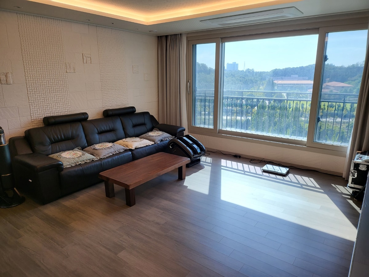 A spacious living room is presented with a large black sectional sofa, complemented by a wooden coffee table. Natural light floods through expansive windows, offering views of the surrounding area. Subtle textures on the walls add to the room's modern design.