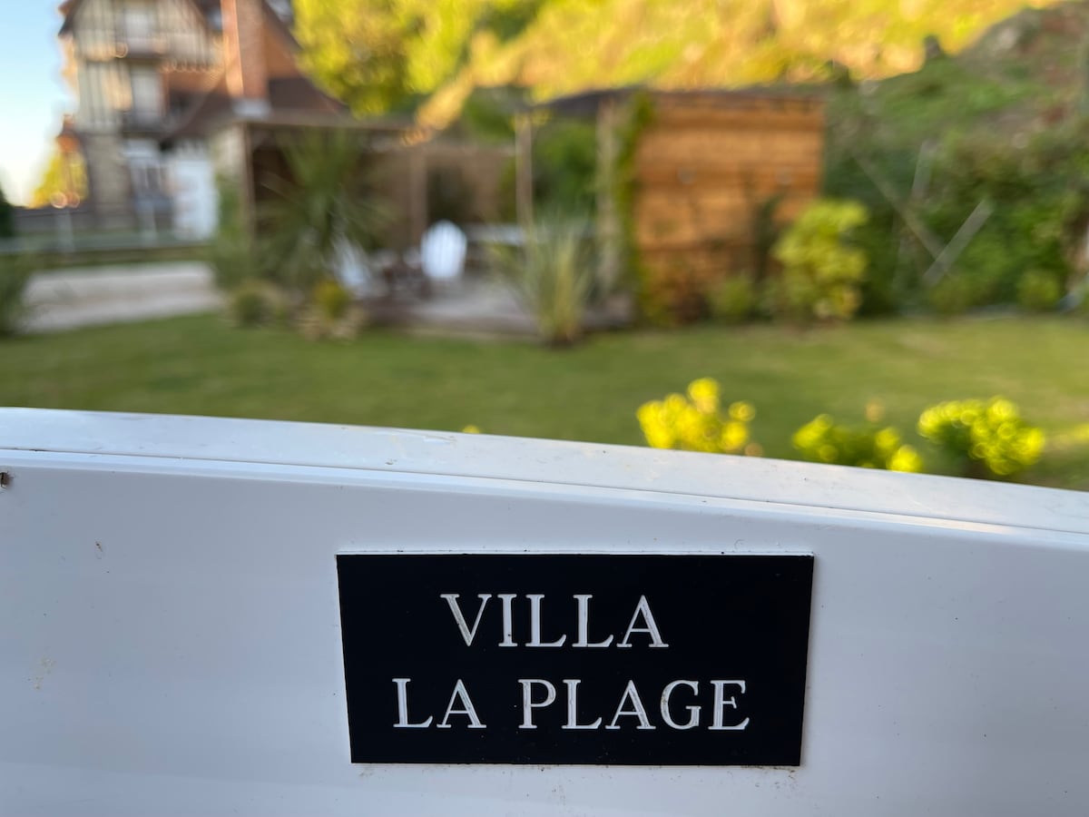 A sign reading 'VILLA LA PLAGE' is displayed prominently. In the background, a landscaped garden with greenery is visible, complemented by a wooden structure, suggesting a peaceful outdoor environment.