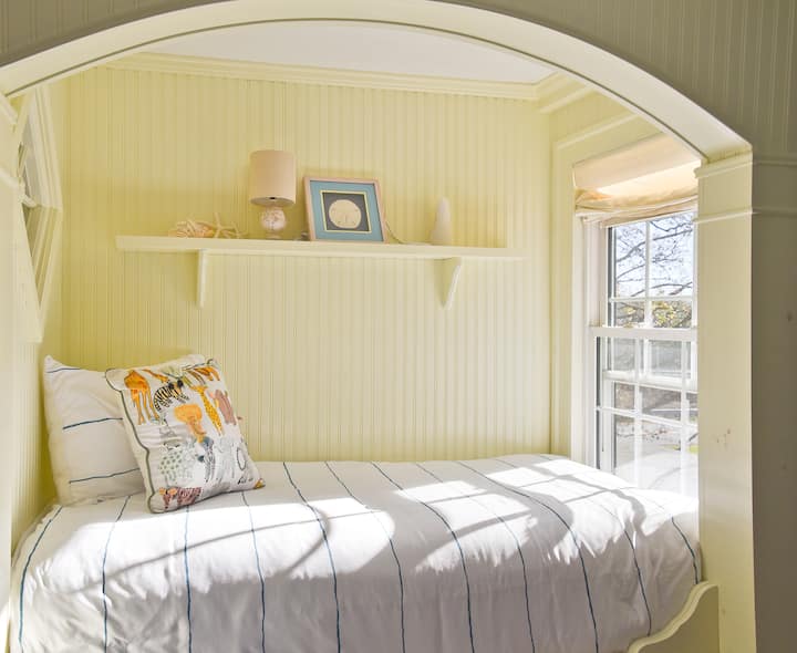 The Captain's Room features a single twin bed built into a custom wood-framed captain's bed with drawers below, a comfortable antique chair, and a pine dresser. 