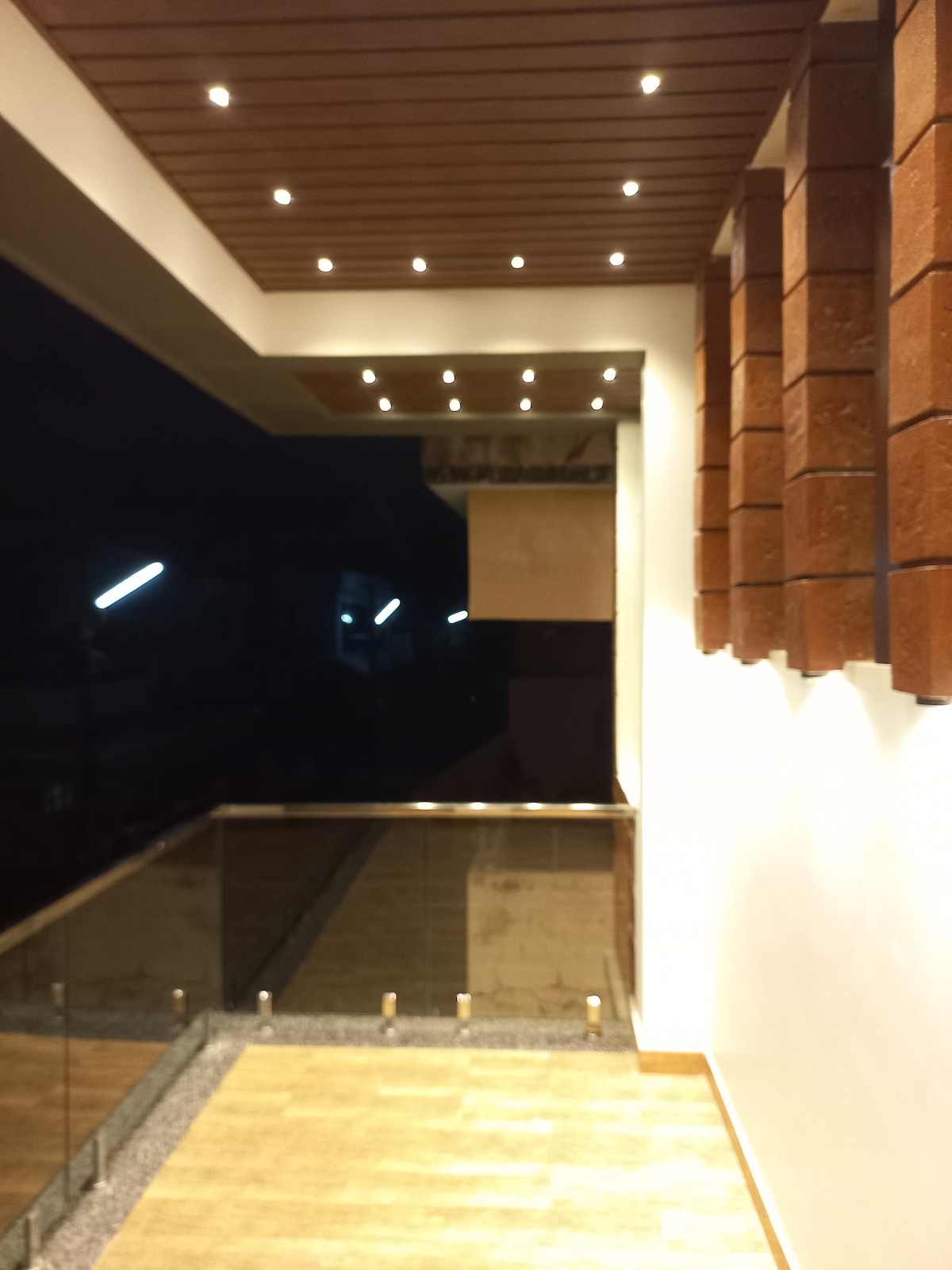 A balcony area features modern wooden ceiling beams with recessed lighting. A glass railing provides an unobstructed view, complementing the natural stone flooring. The scene is illuminated by soft ambient light, creating a peaceful atmosphere suitable for relaxation.