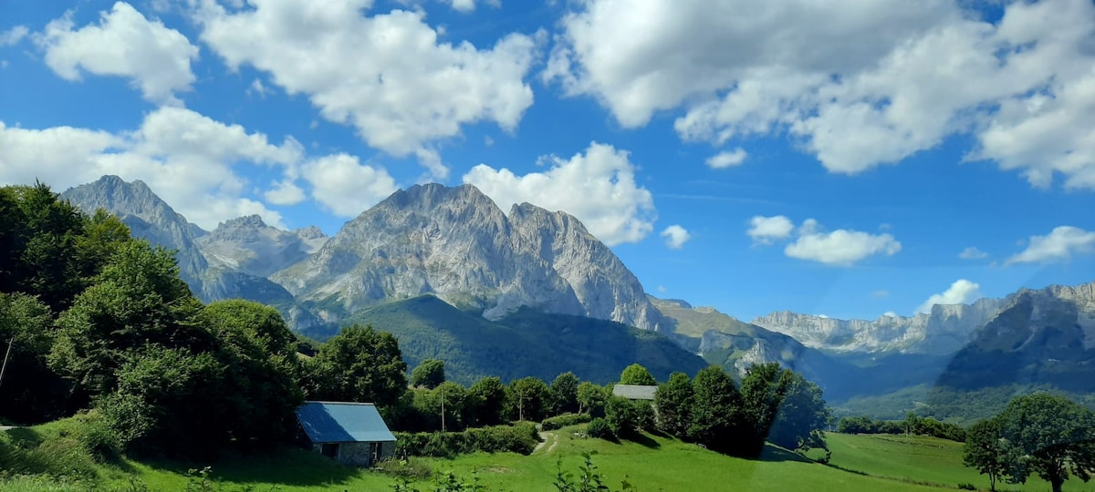 A wide view of the majestic mountains stands under a partly cloudy sky, with lush green fields in the foreground. A small structure is nestled among trees, complementing the idyllic landscape that offers a sense of tranquility and connection to nature.