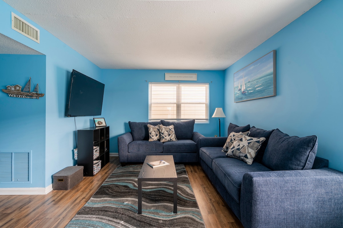 A modern living area displays a comfortable navy blue sectional sofa adorned with decorative pillows. A small coffee table sits in front, with a light area rug beneath. A wall-mounted television is visible, alongside a window allowing natural light to enter the space.