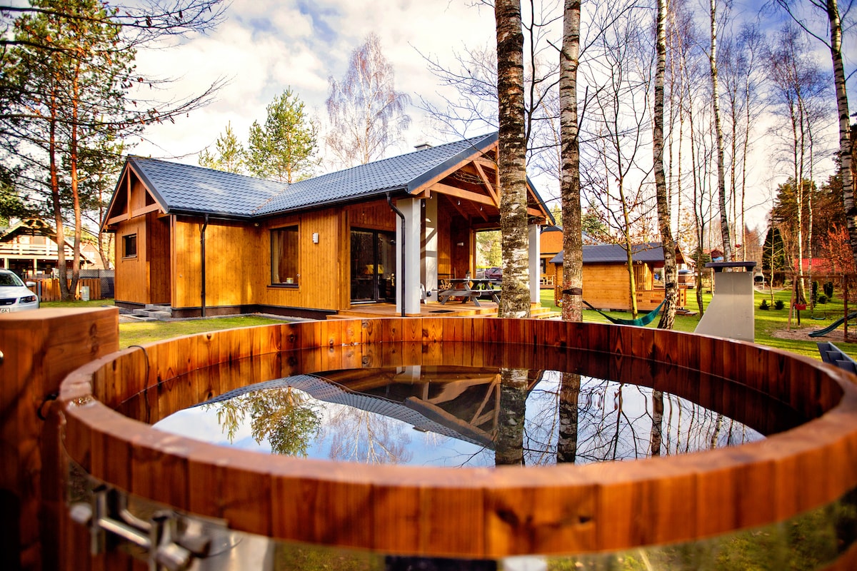 A spacious wooden house is visible, surrounded by trees and greenery under a blue sky. In the foreground, a wooden hot tub reflects the surroundings, adding a serene element to the outdoor space.