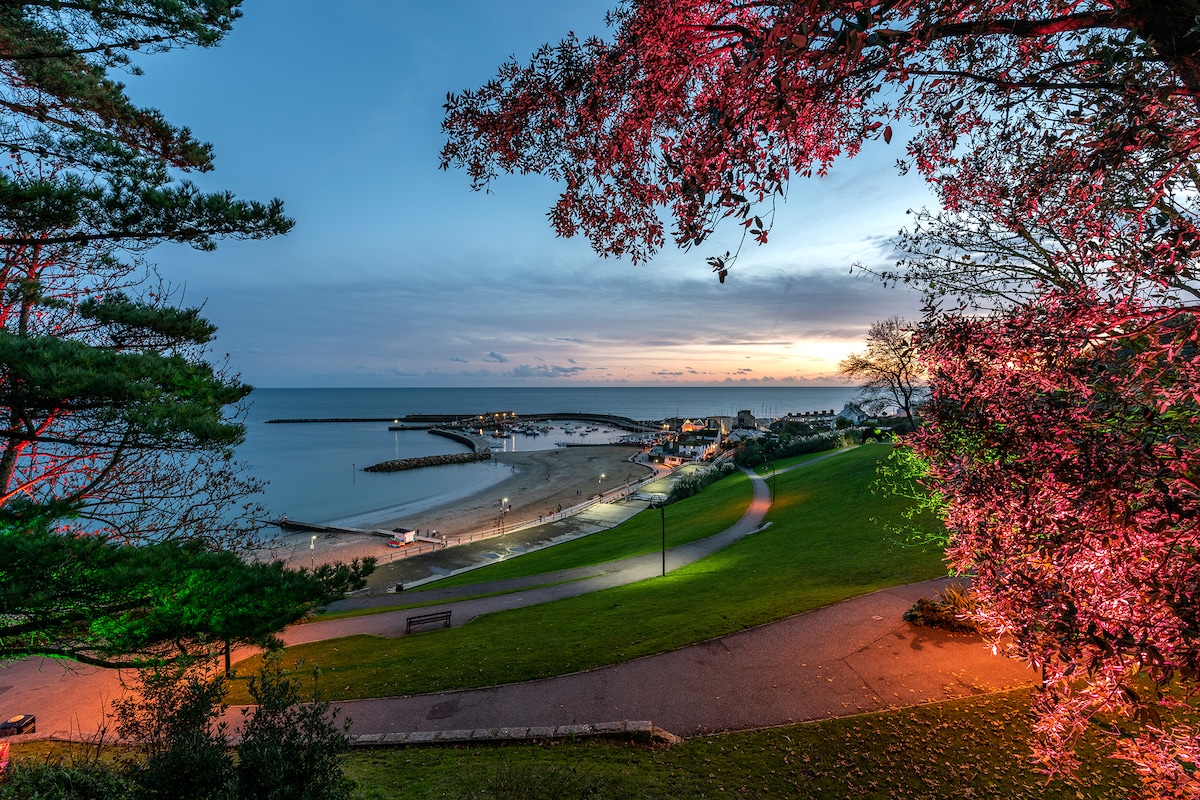 A serene coastal scene at dusk features a calm beach with gentle waves lapping at the shore. A marina with boats is visible in the distance, surrounded by softly illuminated pathways. Lush trees frame the image, enhanced by hues of pink and orange in the sky.