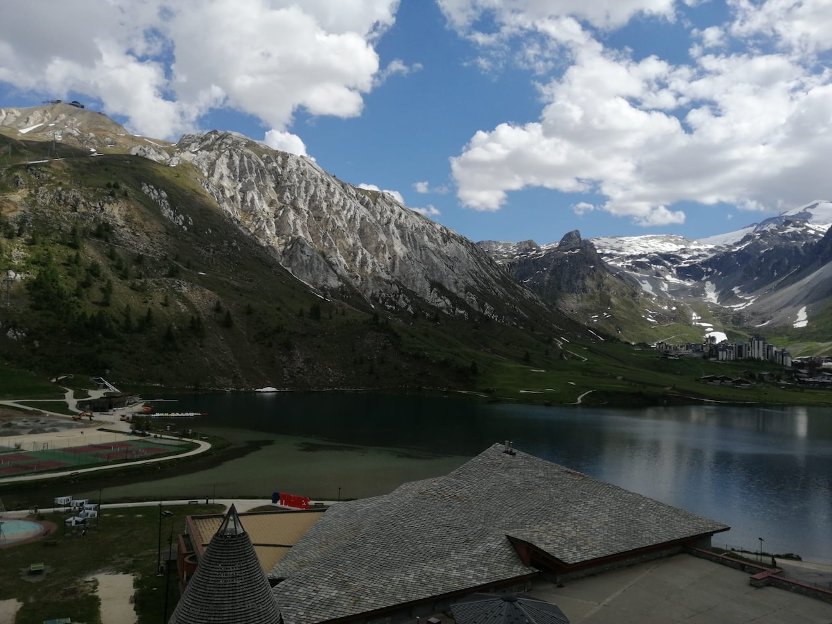 A panoramic view of a serene lake surrounded by rocky mountains and greenery. The reflection of the landscape can be seen in the calm waters, while patches of snow remain on the peaks under a partly cloudy sky.