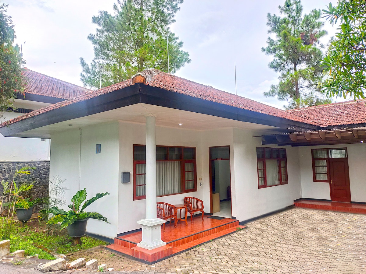 A single-story house presents a covered entry with a red-tiled roof. A small seating area with two wooden chairs and a table is positioned on a tiled porch. Surrounding greenery and tall trees enhance the peaceful exterior. The driveway features patterned stone paving.