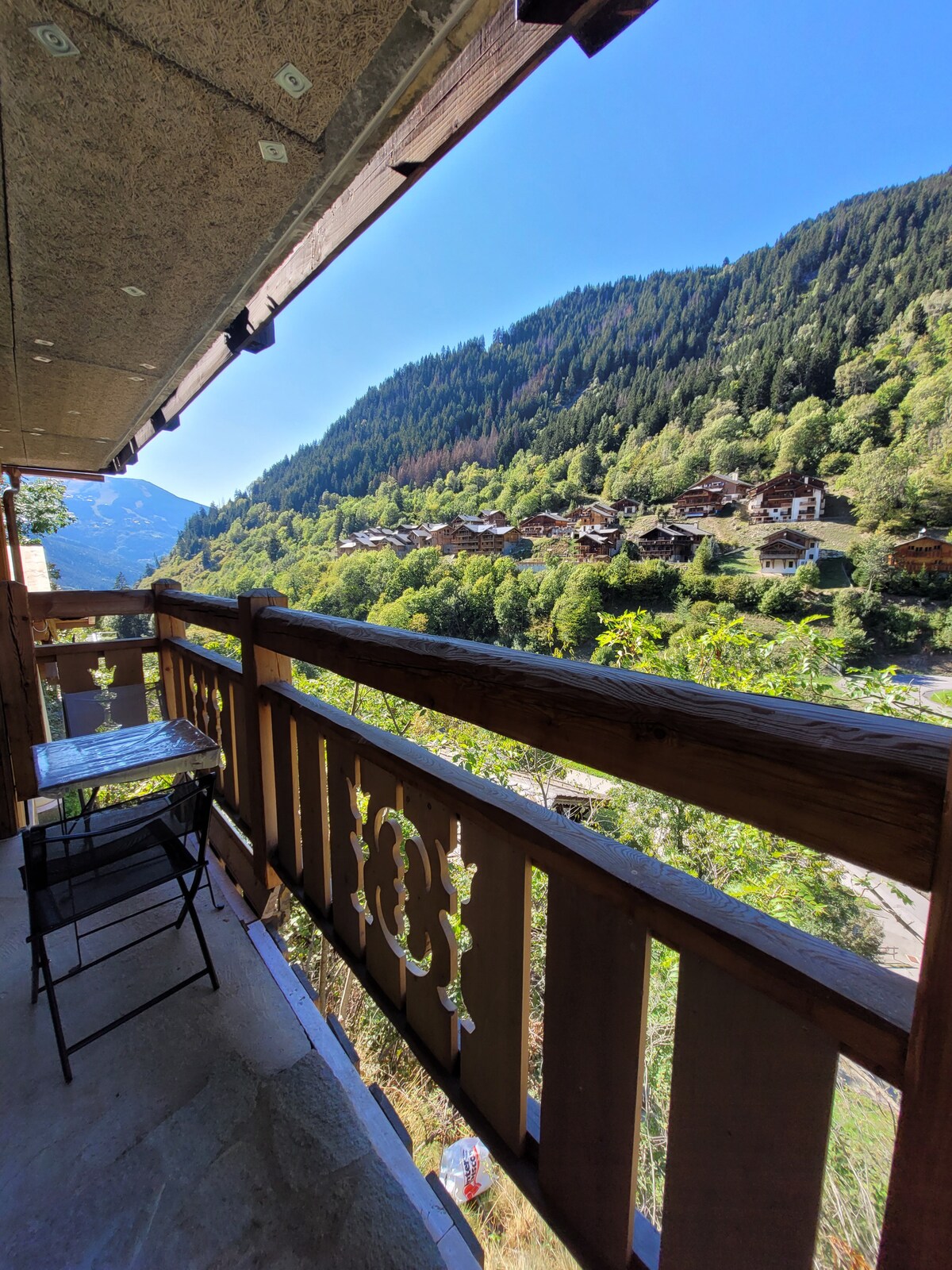 A balcony with a wooden railing offers a serene view of the surrounding mountains and lush greenery. A small table and chair are placed on the balcony, providing a cozy spot to enjoy the scenery. The clear blue sky complements the tranquil atmosphere.