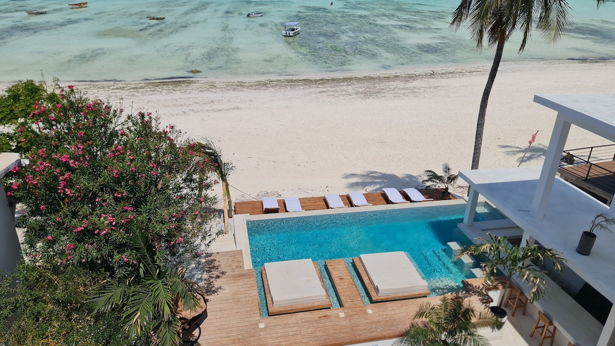 An outdoor area is visible featuring a serene infinity pool adjacent to a sandy beach. Comfortable lounge beds are arranged on a wooden deck, surrounded by lush greenery and tropical flowers. The turquoise water of the ocean creates a calming backdrop against the clear sky.