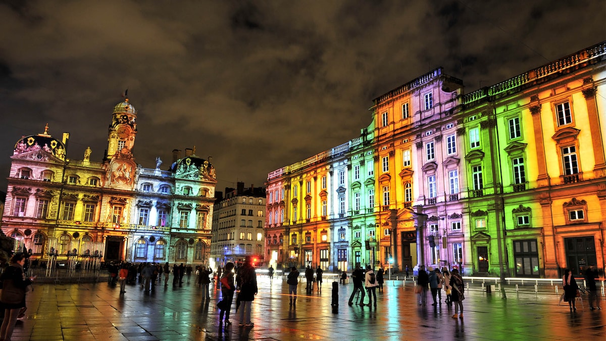 The illuminated Place des Terreaux features historic buildings with vibrant light projections, creating a colorful atmosphere at night. Crowds of people are seen gathering, with reflections visible on the wet cobblestone surface, enhancing the lively ambiance of the square.