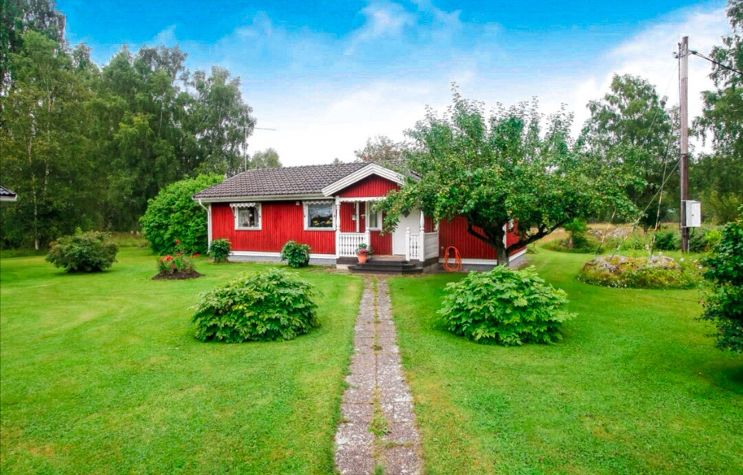 A charming red house is surrounded by a lush green garden. A stone pathway leads to the entrance, flanked by neatly trimmed bushes and trees. The exterior features a covered porch and large windows that invite natural light.