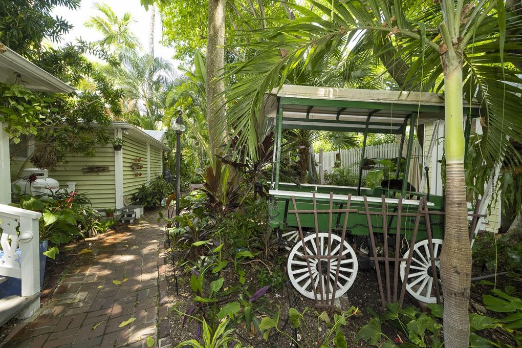 A lush garden pathway is surrounded by tropical plants and palm trees, creating a serene environment. A vintage green cart is positioned along the path, complementing the natural beauty with its rustic charm. Soft dappled light filters through the foliage.