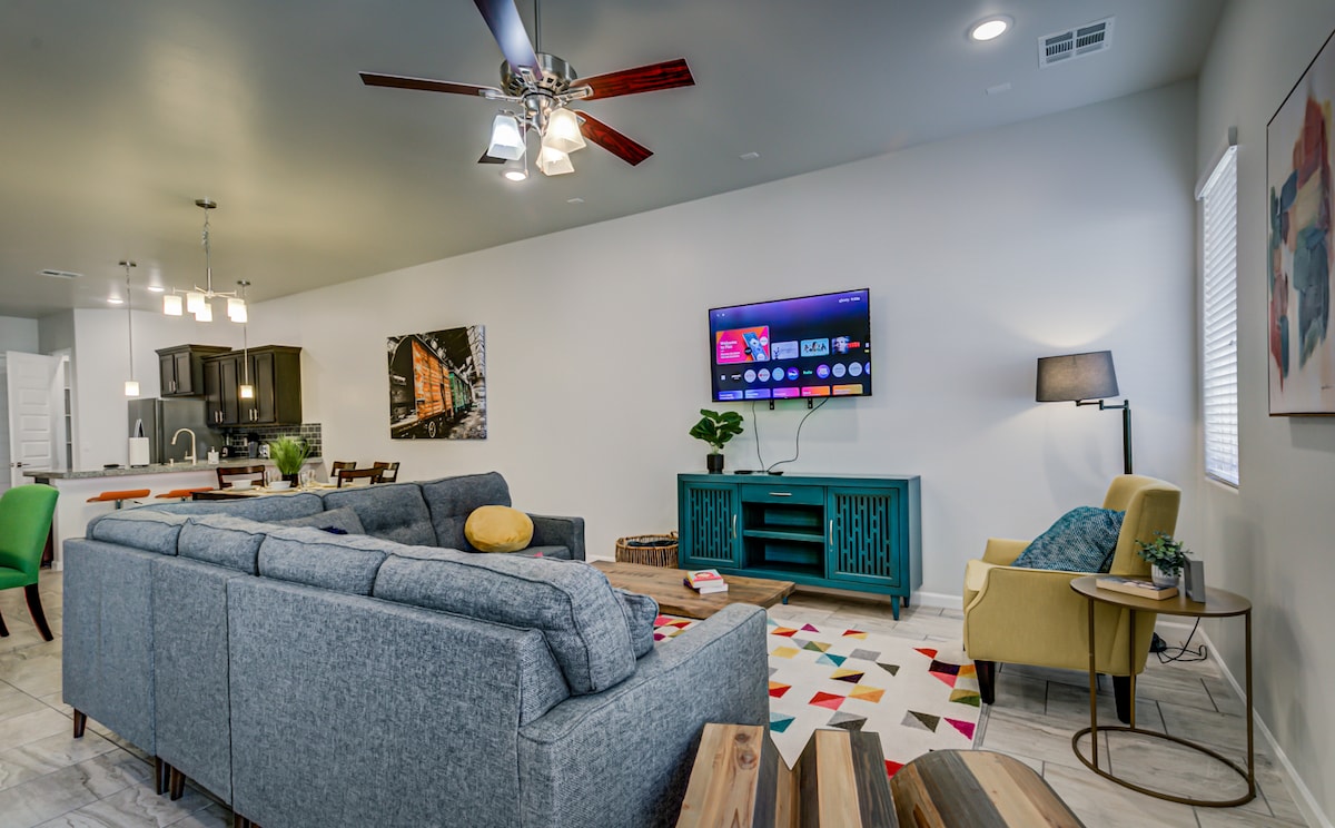 A spacious living area features a large sectional sofa and a colorful area rug. A smart TV is mounted on the wall, and a stylish side table with a lamp is positioned next to a chair. The room includes modern decor and soft lighting.