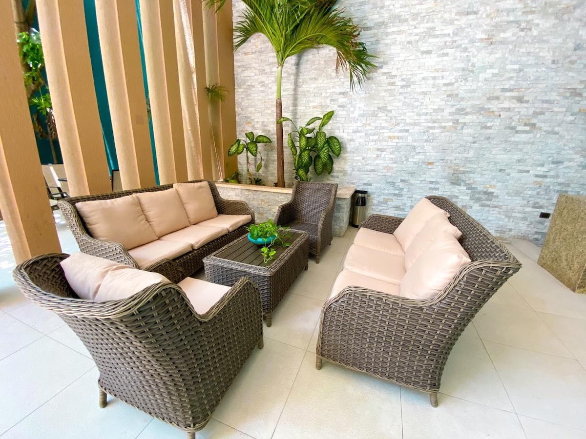 A seating area is displayed with woven furniture, featuring a light-colored upholstered sofa and armchairs arranged around a central coffee table. Lush potted plants and a textured stone wall enhance the space, creating a welcoming environment for relaxation.