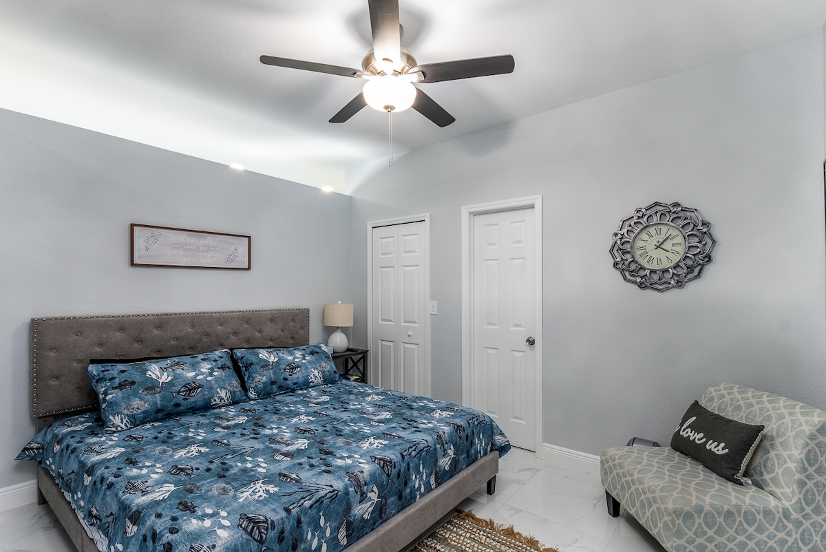 A bedroom is presented with a comfortable king-size bed featuring a blue patterned duvet. A nightstand with a lamp is positioned next to the bed. The decor includes a clock and wall art. A small seating area is available, enhancing the room's functionality.