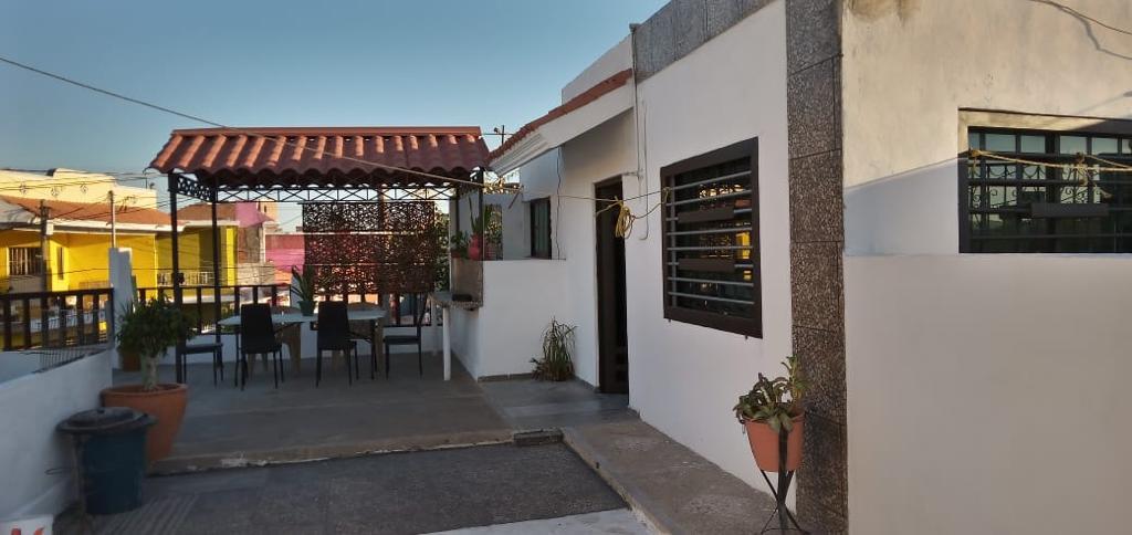 An outdoor terrace area is visible, featuring a covered seating arrangement with a table and six chairs. Surrounding plants and a small potted tree add greenery to the space. The terrace provides a view of the surrounding neighborhood under a clear sky.