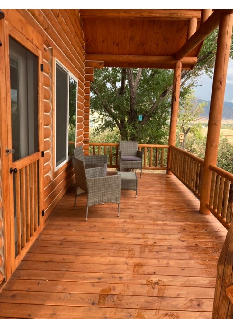 An outdoor deck features wooden flooring and log railings, showcasing two comfortable chairs positioned beside a large window. The surrounding trees add natural greenery, while the spacious area offers ample room for relaxation and enjoying the mountain views.