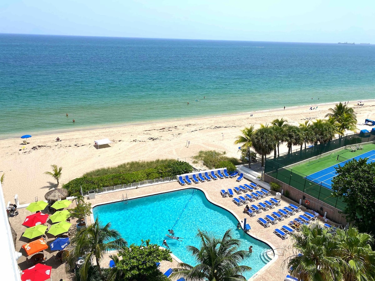 A view of a sparkling pool is framed by tropical palm trees and surrounded by lounge chairs. Beyond the pool, the sandy beach meets the inviting blue ocean, with beach umbrellas set up along the shore. The scene conveys a vibrant outdoor atmosphere.