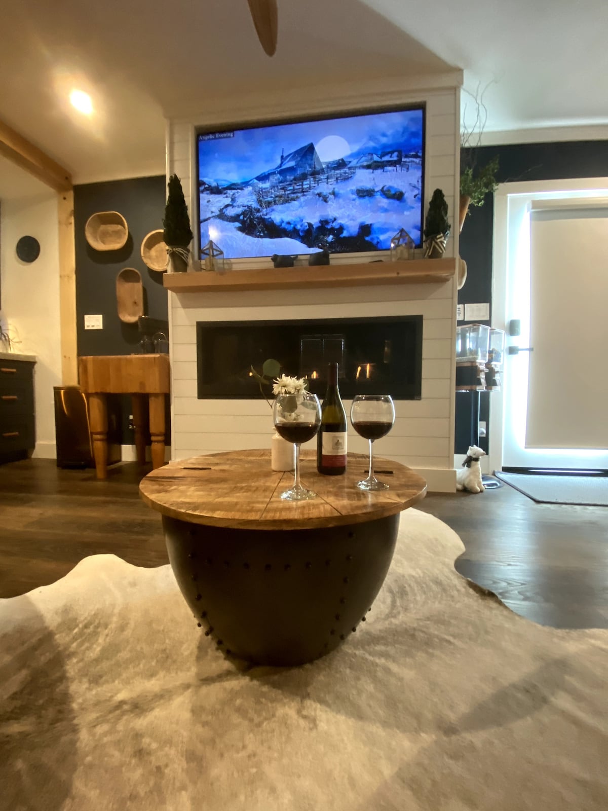 A cozy living area is highlighted by a wooden coffee table with two glasses of wine. A large television displays a scenic mountain view. A fireplace is positioned beneath the TV, contributing to the inviting ambiance. The space features a soft area rug and warm, neutral tones.