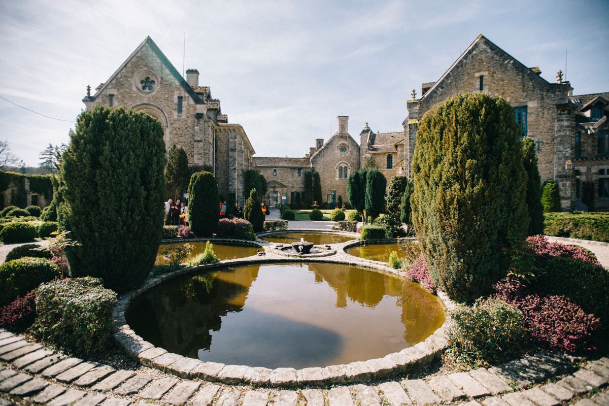 A serene courtyard is framed by well-manicured hedges and various shrubs, surrounding a reflective pond. The historic stone buildings feature distinct architecture, with large windows and intricate details. Bright sunlight illuminates the scene, accentuating the peaceful atmosphere of the surroundings.
