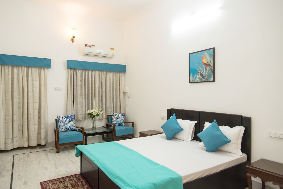 A spacious bedroom features a large bed with a turquoise bedspread and white pillows. Two comfortable chairs with vibrant cushions are arranged beside a small table. Natural light enters through windows covered with light curtains, enhancing the serene ambiance.