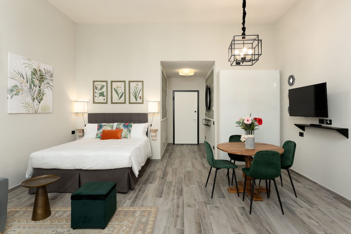 A spacious studio features a comfortable bed with decorative pillows and a seating area with a small green chair and a dark ottoman. A dining table with green chairs is set against a bright wall, complemented by framed botanical art and a mounted television.