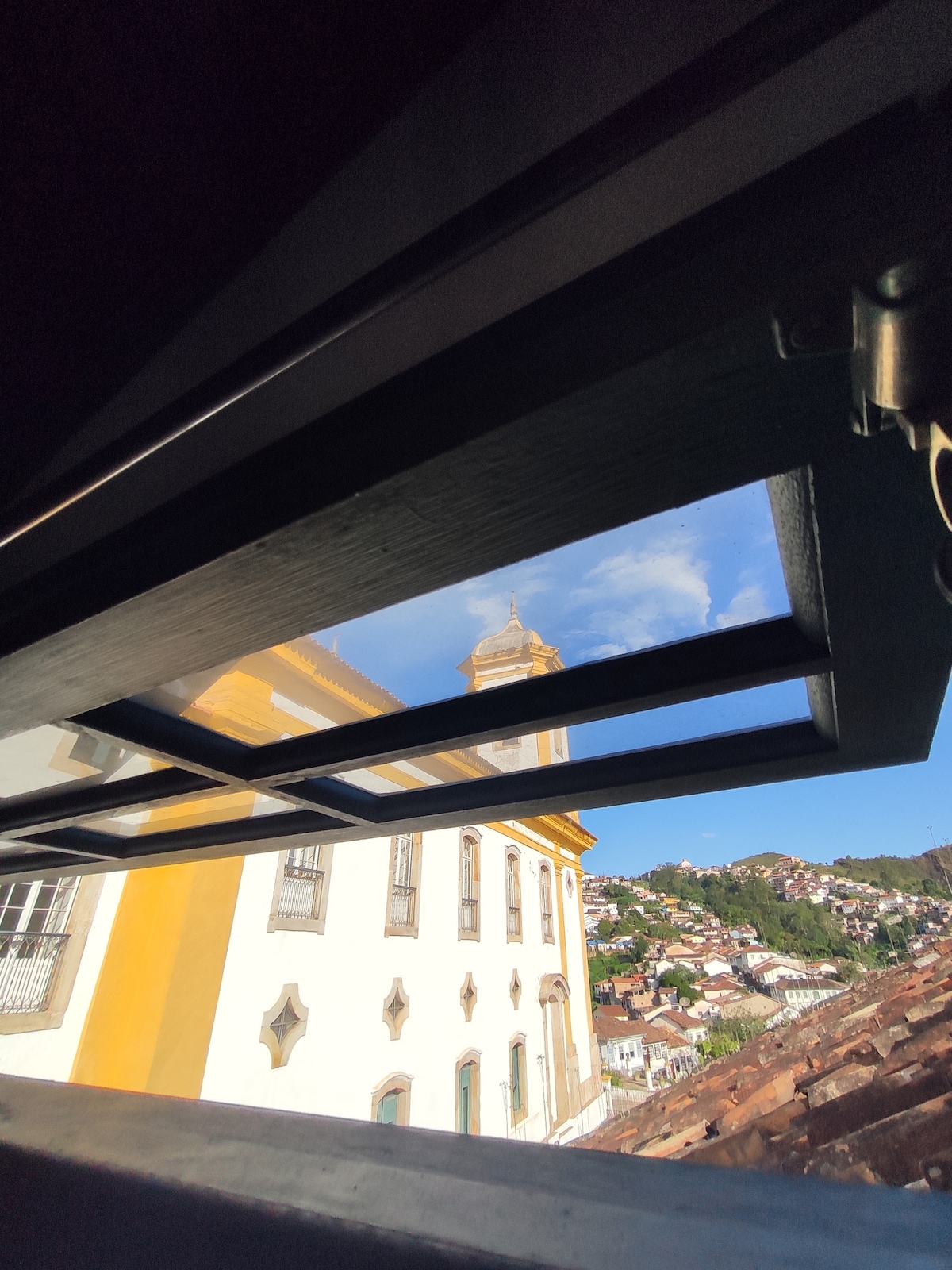 A view from a window showcases the exterior of a historic building, featuring a yellow facade and a distinctive clock tower. The surrounding rooftops and hills of Ouro Preto are visible, with clear blue skies enhancing the scene.