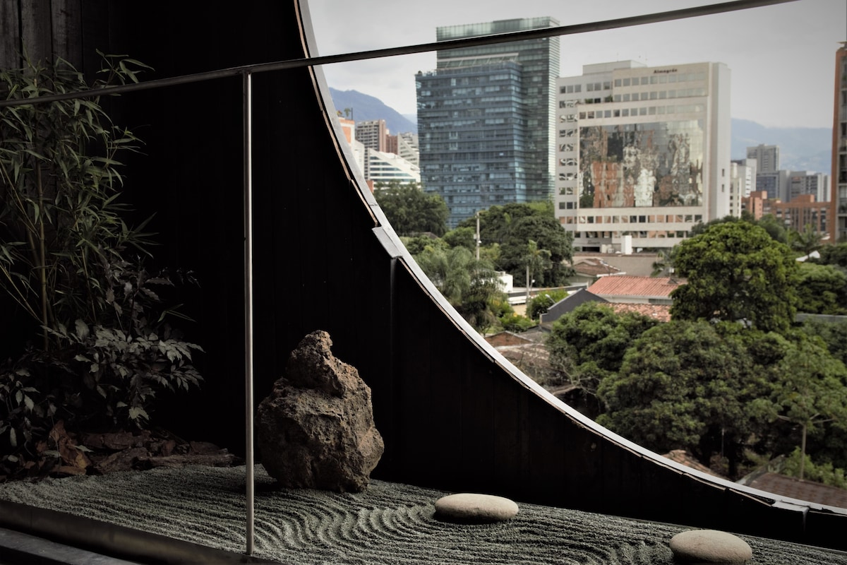 A tranquil outdoor space features a curved balcony with a modern railing. The scene includes a stone and bamboo accent, complemented by a view of Medellin’s skyline, showcasing both greenery and city buildings in the background.