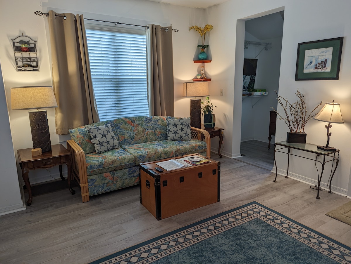 A cozy living area features a floral-patterned sofa, complemented by two decorative lamps and a wooden coffee table. Natural light filters through a window, enhancing the room's inviting atmosphere. A console table and an accent table with a decorative arrangement provide additional charm.
