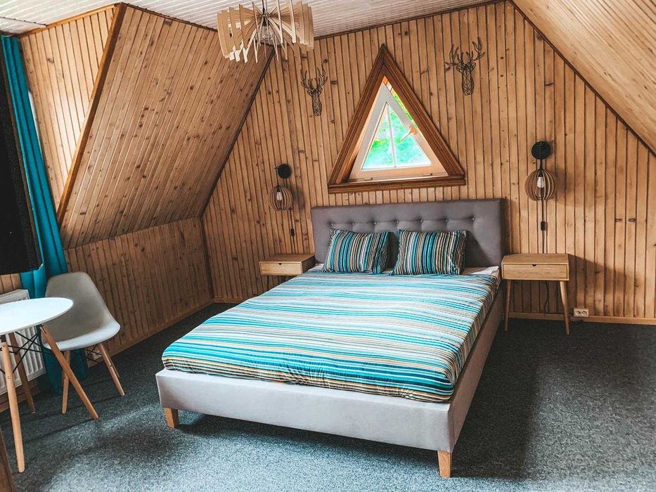 An attic bedroom features wooden paneling and triangular windows, allowing natural light to filter through. A spacious bed with a striped blue and green comforter is positioned centrally, flanked by two light wood nightstands. A small round table is located near the window.
