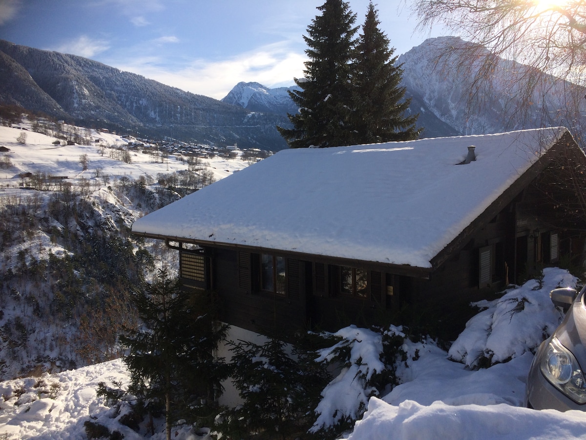 The chalet is surrounded by a snowy landscape, showcasing a wooden exterior with large windows. Tall evergreen trees frame the structure, while distant mountains provide a stunning backdrop. A clear blue sky adds brightness to this tranquil, wintry scene.