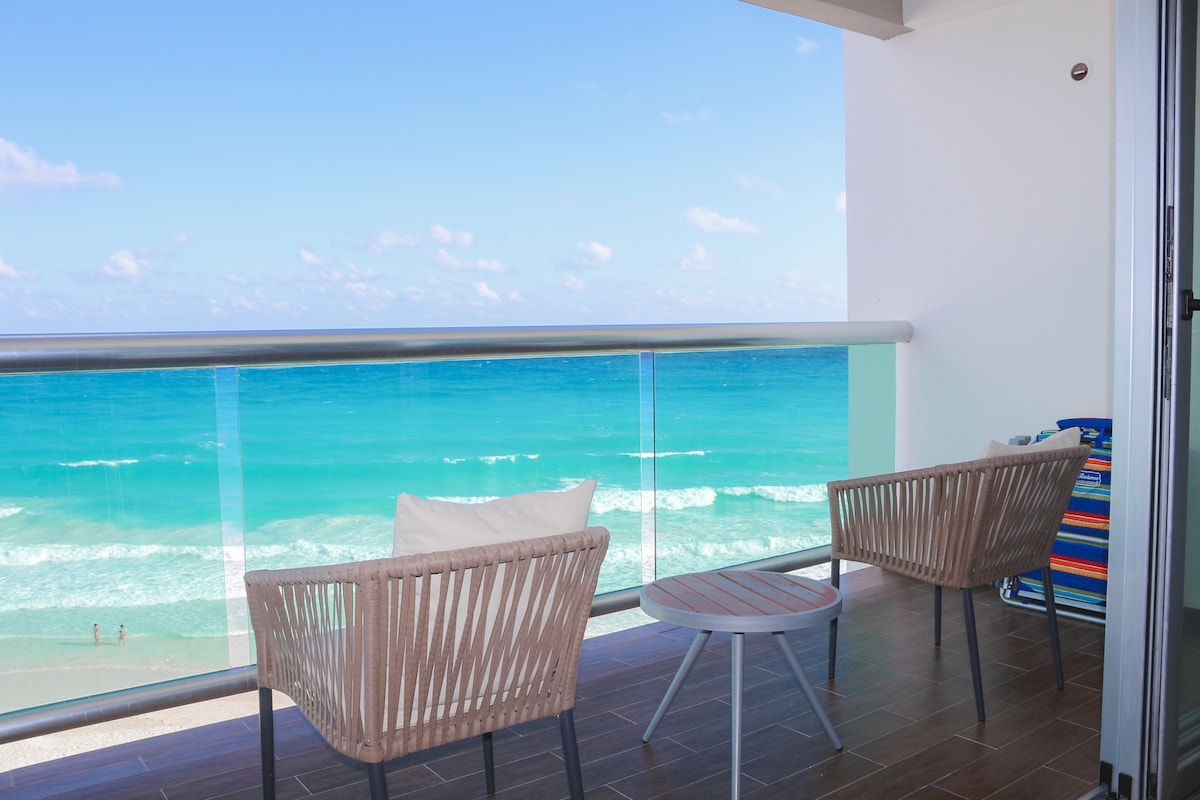 A private terrace offers stunning ocean views, with two modern armchairs and a small table positioned to maximize the scenery. The turquoise water can be seen gently lapping against the shore, complemented by a clear blue sky dotted with soft clouds.