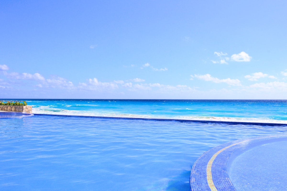 A tranquil infinity pool extends towards the horizon, blending seamlessly with the vibrant blue ocean. Soft waves can be seen lapping against the shore in the distance, under a clear sky dotted with light clouds.
