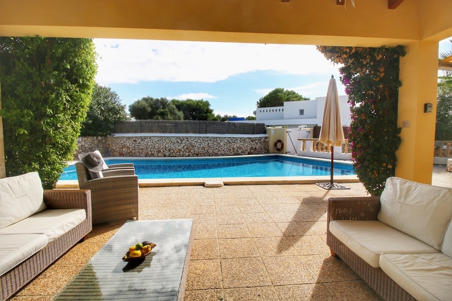 A covered patio area is depicted, featuring two light-colored sofas and a glass coffee table. The inviting swimming pool is visible in the background, framed by greenery and surrounded by a stone wall. Sunlight brightens the space, creating a relaxed outdoor atmosphere.